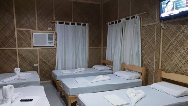 Habitación con cuatro camas individuales, cada una con ropa blanca y toallas, un televisor montado en la pared y un equipo de aire acondicionado.
