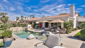 Un área de piscina con sillones de descanso, un jacuzzi y una pérgola con sombrillas.