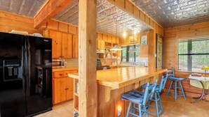 A kitchen with wooden cabinets, a black refrigerator, a bar area with blue stools, and a window with blinds. - Spacious one bedroom log cabin with views for miles! Only one mile from the Wimb (Wimberley)