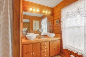 Une salle de bain de style rustique, avec des murs en bois, un meuble-lavabo, deux éviers, un miroir et une fenêtre ornée de rideaux en dentelle.