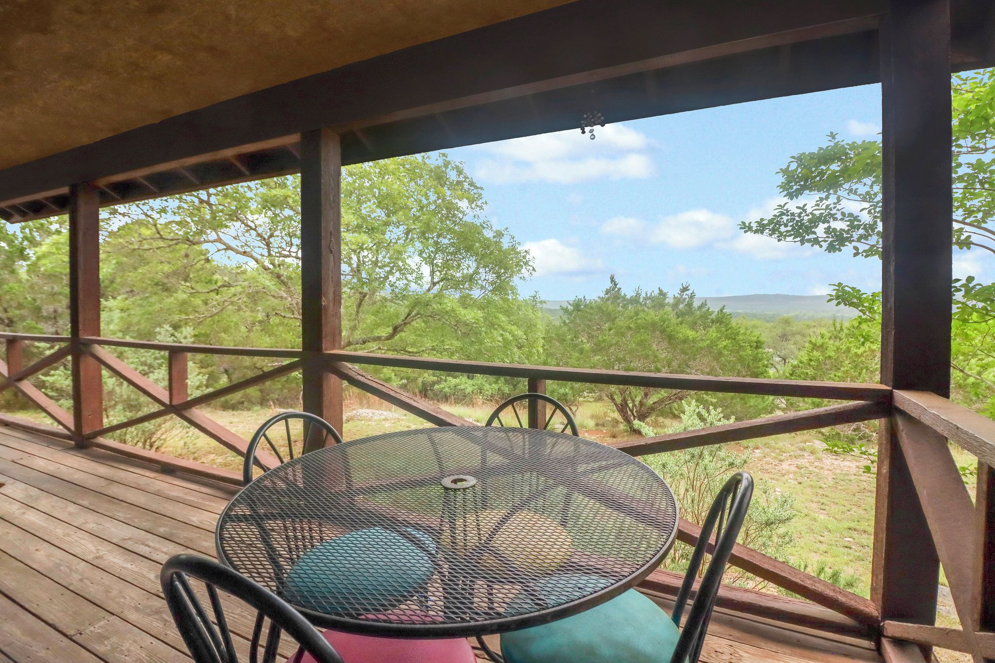A wooden deck with a metal table and chairs, overlooking a natural landscape with trees and hills.