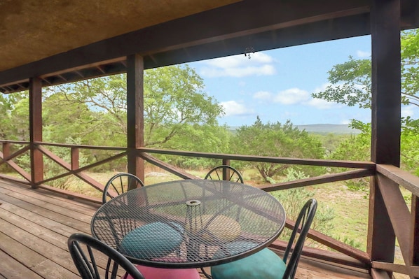 A wooden deck with a metal table and chairs, overlooking a natural landscape with trees and hills.