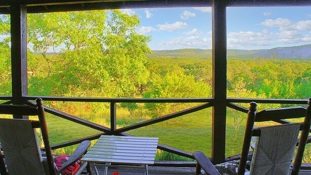 A covered outdoor seating area with a table and chairs, overlooking a lush landscape with trees and hills. - Spacious one bedroom log cabin with views for miles! Only one mile from the Wimb (Wimberley)