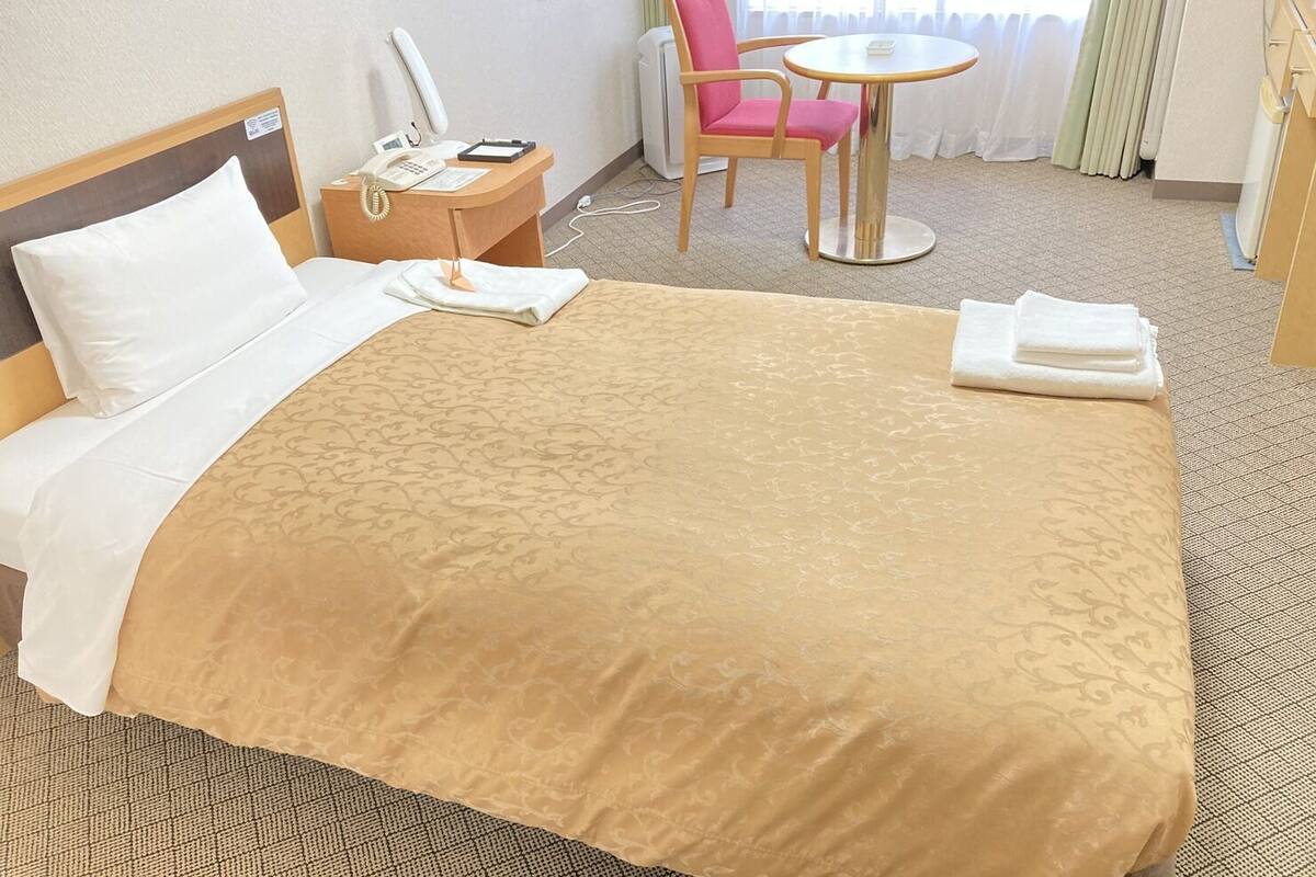 A hotel room with a bed, a bedside table with a phone and a lamp, a chair, a small round table, and a window with curtains.