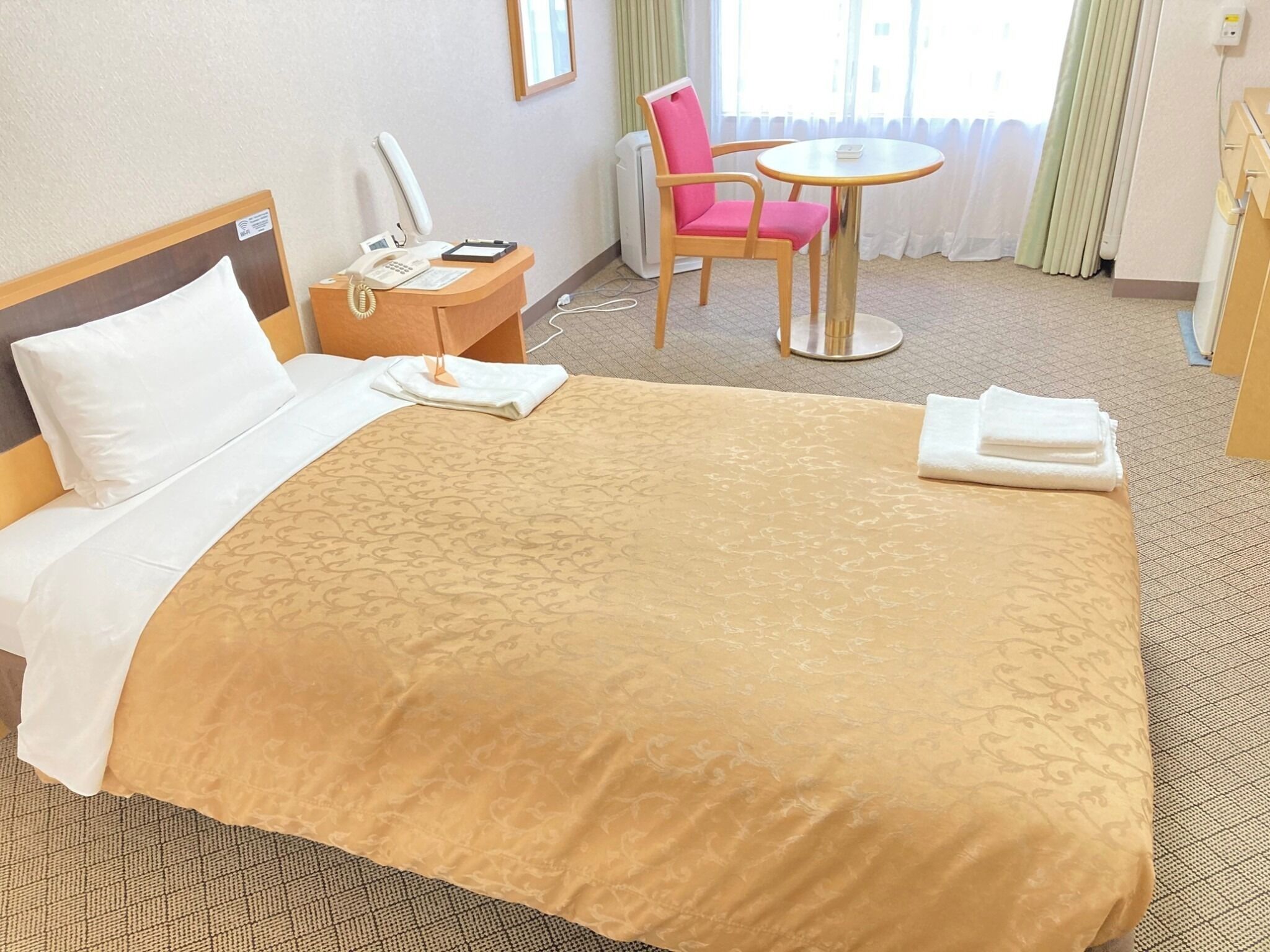 A hotel room with a bed, a bedside table with a phone and a lamp, a chair, a small round table, and a window with curtains.