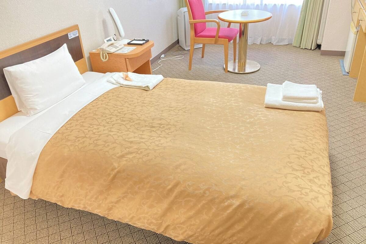 A hotel room with a bed, a bedside table with a phone and a lamp, a chair, a small round table, and a window with curtains.