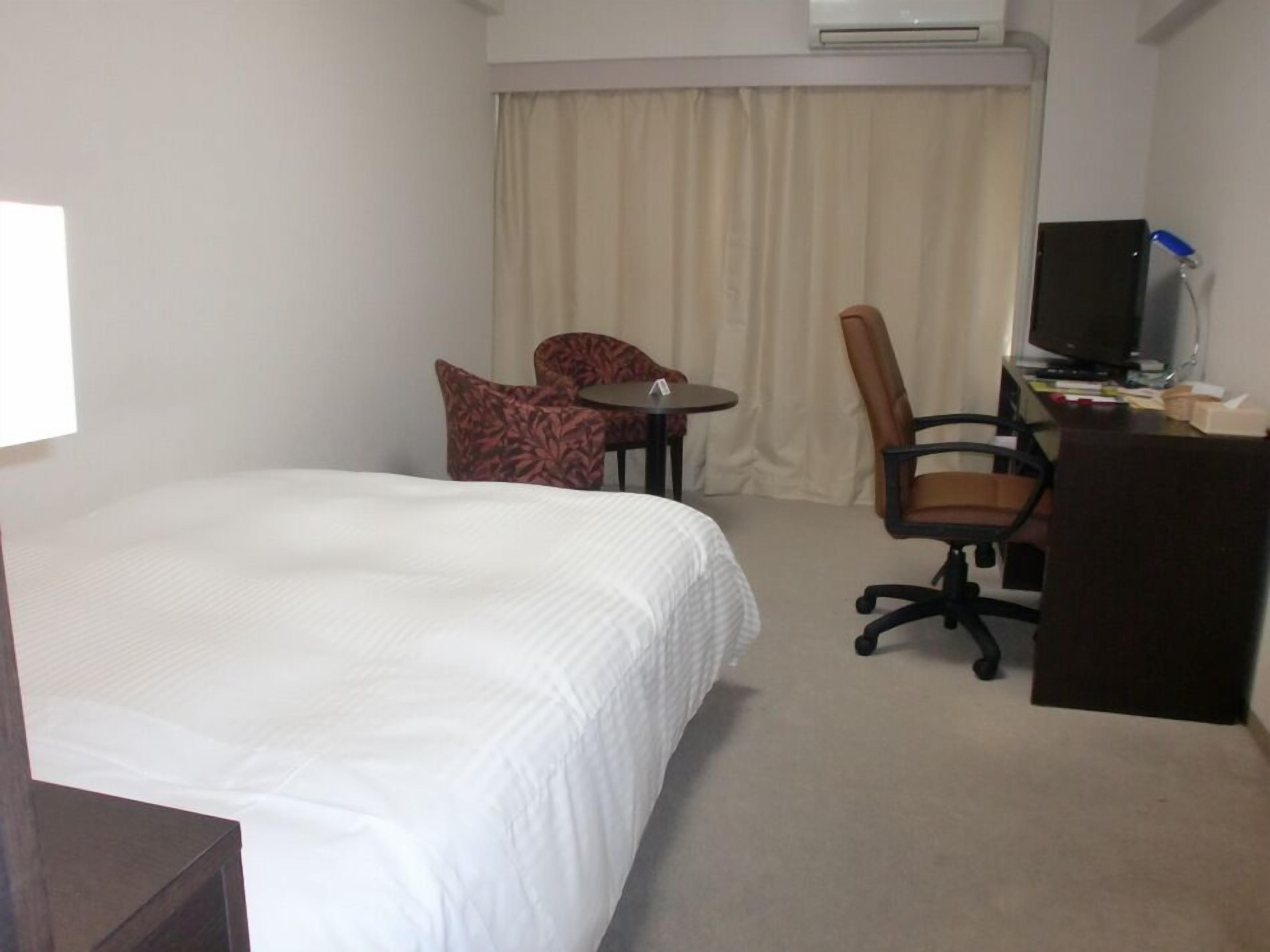 A hotel room with a bed, a desk, a chair, a television, and an air conditioning unit.