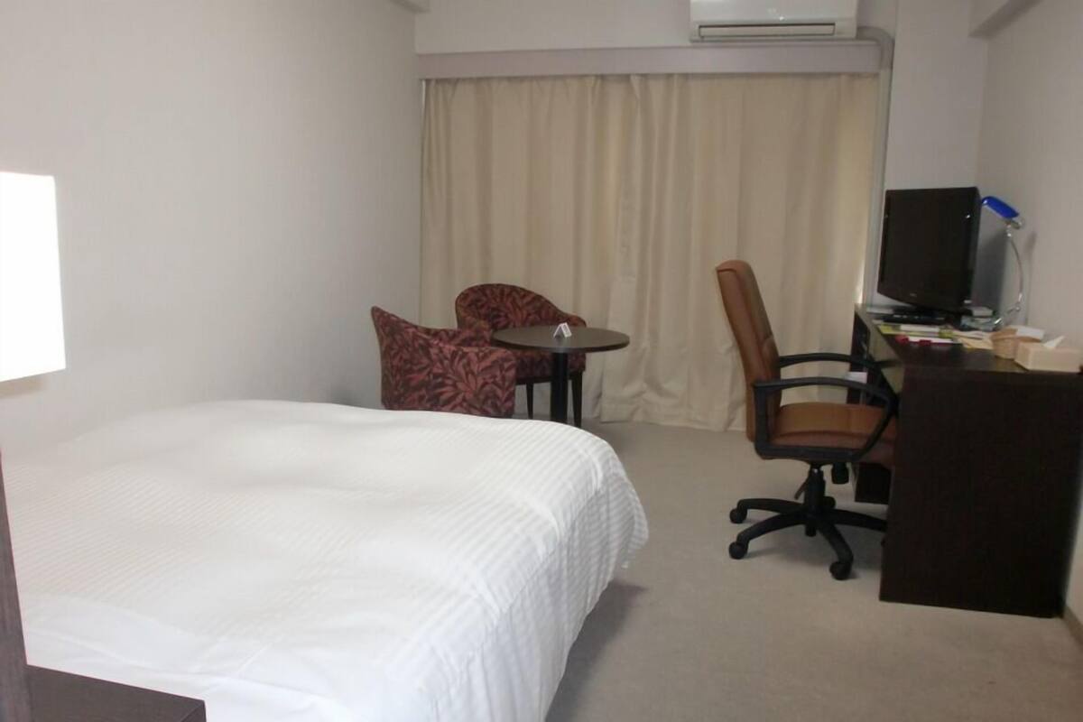 A hotel room with a bed, a desk, a chair, a television, and an air conditioning unit.