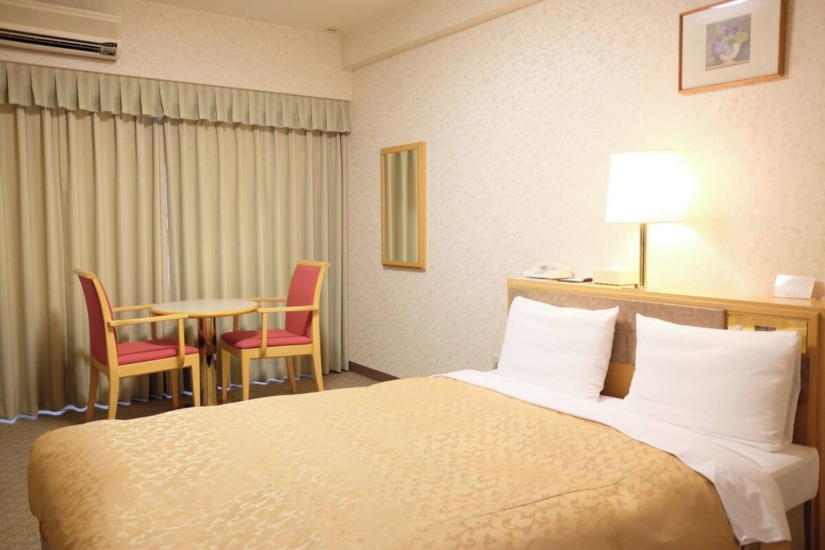 A hotel room with a bed, a desk with a chair, a lamp, and a mirror on the wall.