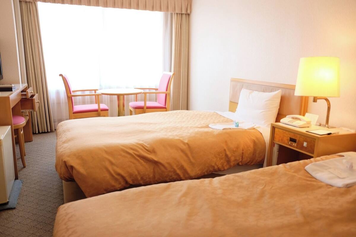 A hotel room with two beds, a desk, a chair, a lamp, and a window with curtains.