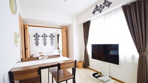 A hotel room with a wooden desk, a chair, a flat-screen TV, and a large window with curtains.
