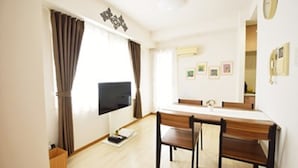 A modern living room with a flat-screen TV mounted on the wall, a dining table with chairs, and a large window with curtains.