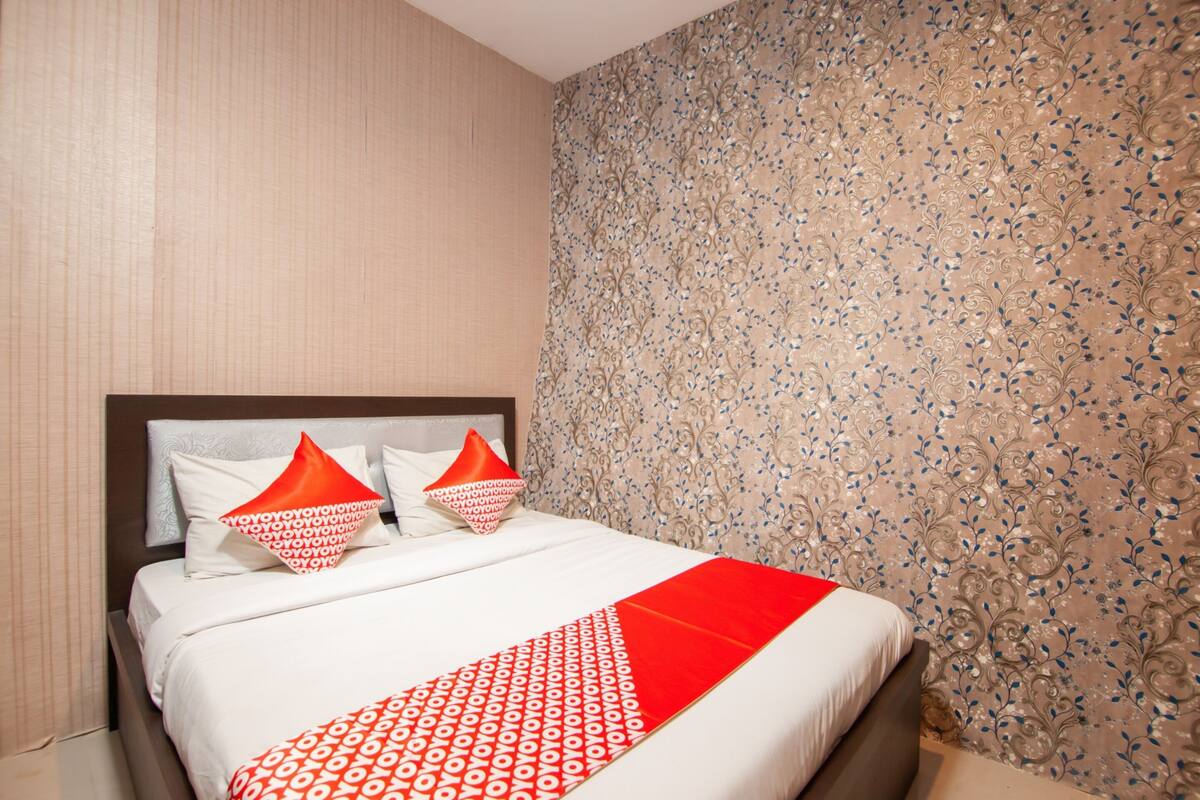 Quarto de hotel com uma cama, duas almofadas, edredom estampado em vermelho e branco, e papel de parede floral na parede.