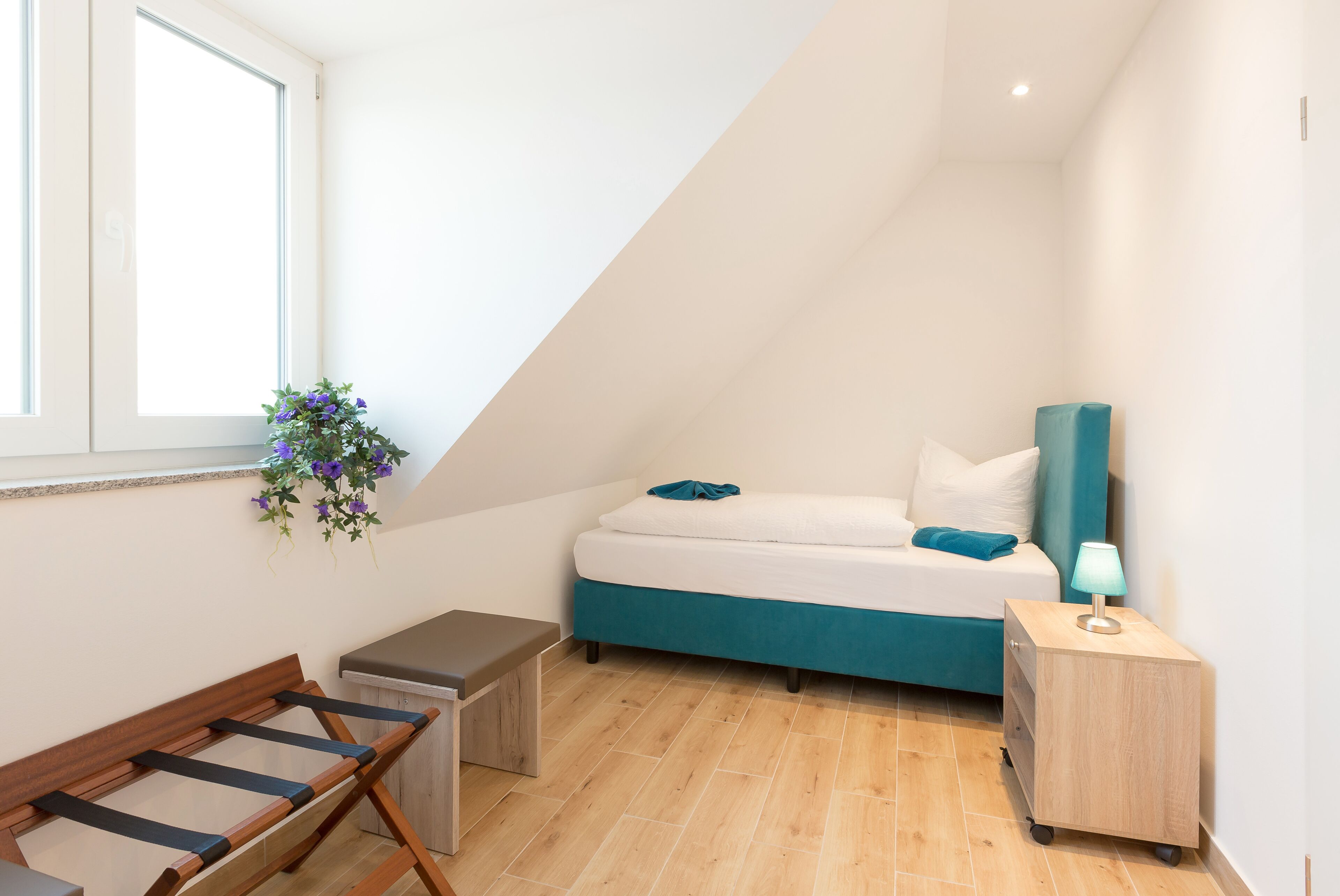 Quarto pequeno e moderno com cama, um banquinho, uma escrivaninha e uma janela com uma planta.