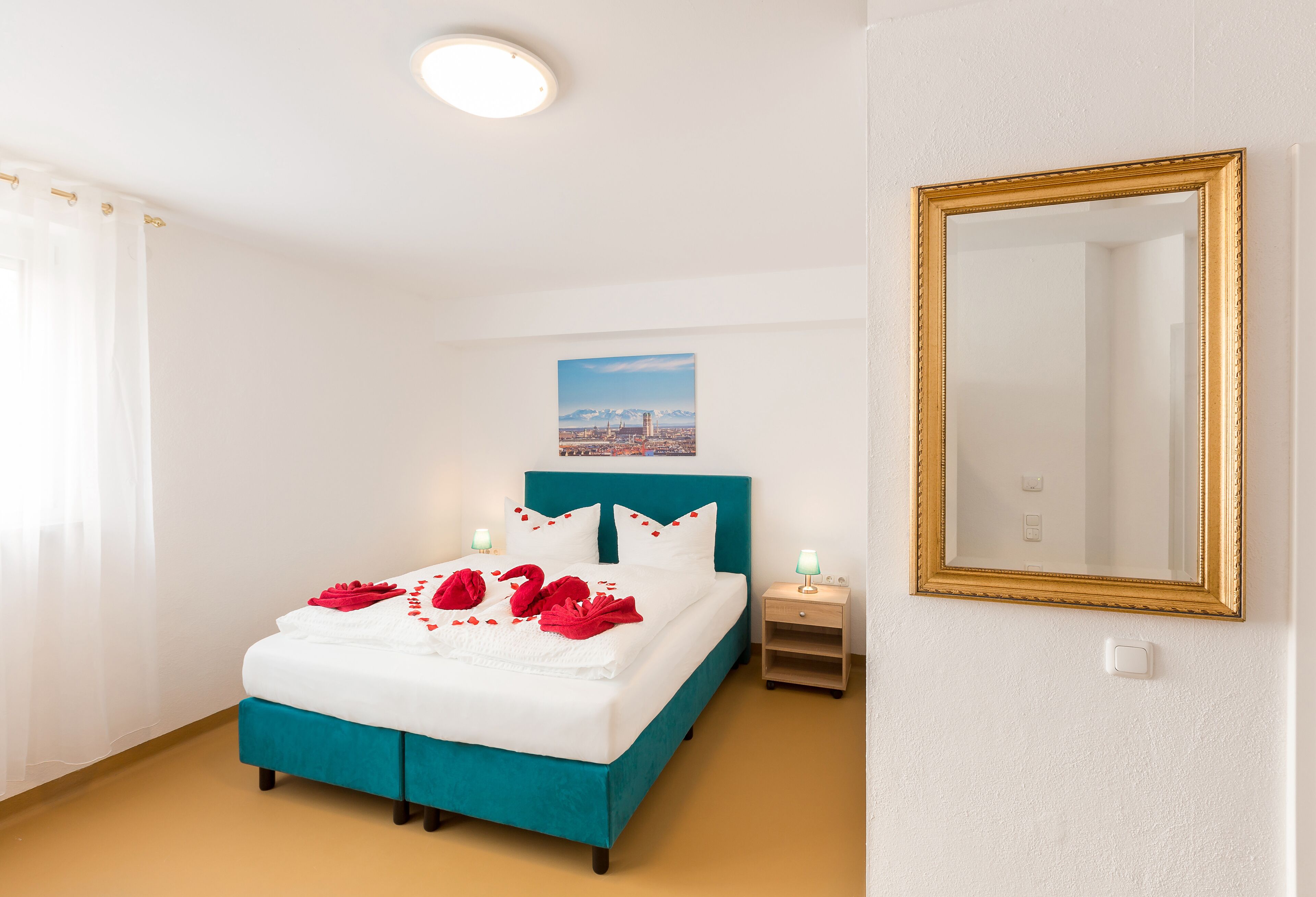 A neatly made bed with a teal headboard, white bedding, and red accents. There is a framed mirror on the wall and a small bedside table with a lamp.
