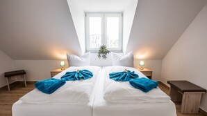 A double bed with white linens and teal bedding, two bedside tables with lamps, a bench, and a window with a view of buildings. - Hotel Ludwig (Munich)