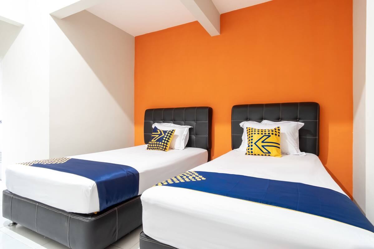 A hotel room with two beds, each with a black headboard and white bedding. The walls are orange, and there is a white ceiling with a skylight.