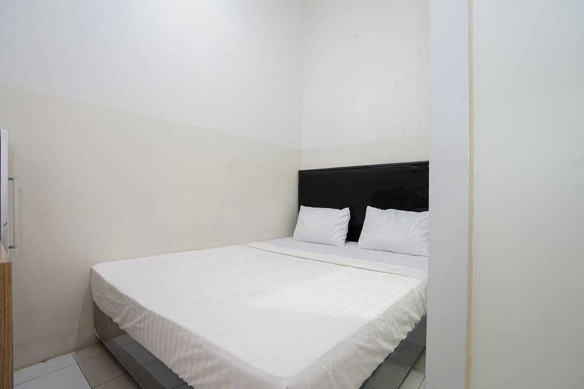 Quarto pequeno e limpo, com uma cama de solteiro, paredes brancas e uma porta de madeira simples.