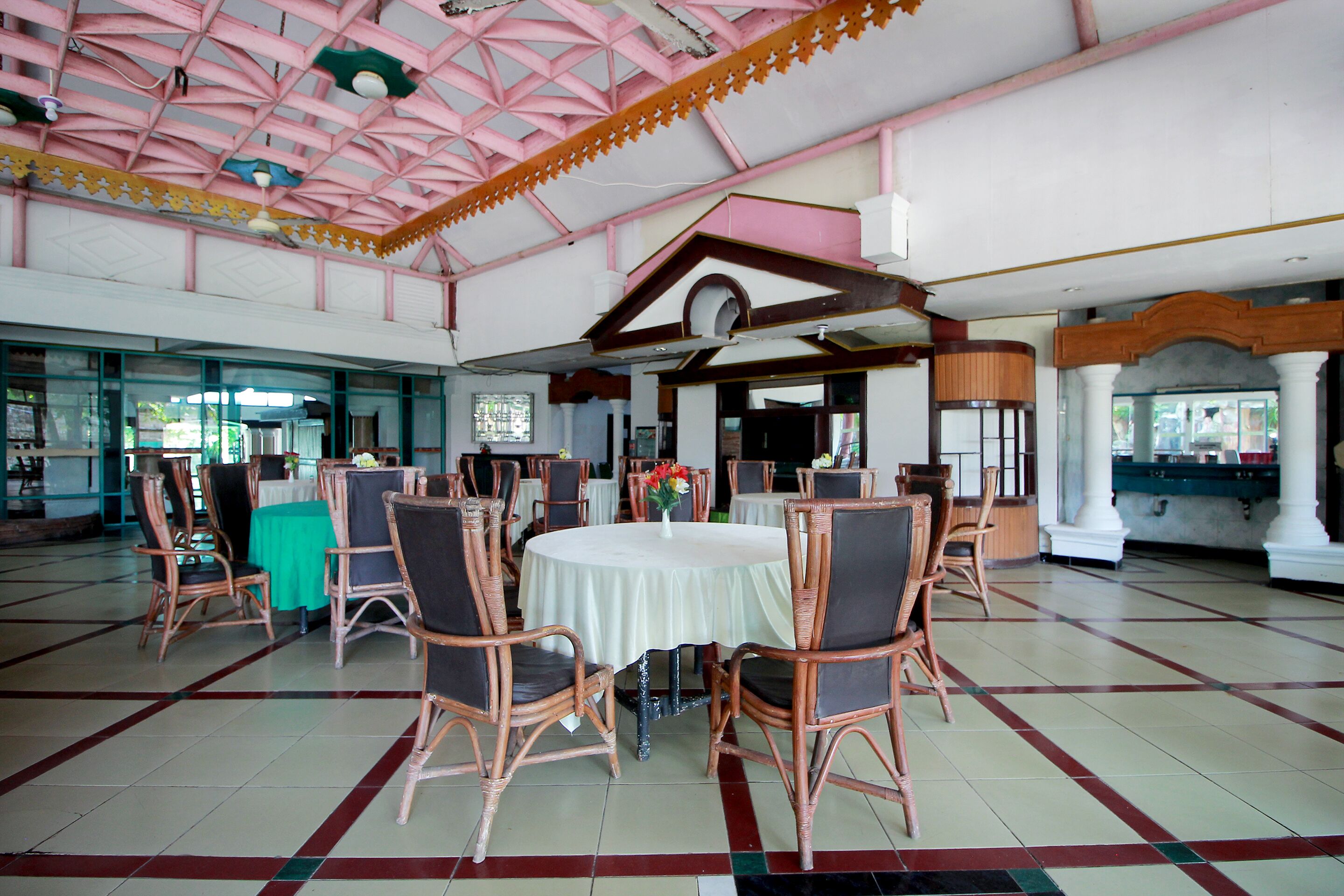A spacious hall with round tables and wicker chairs, a high ceiling with a geometric pattern, and a view of the outside through large windows.