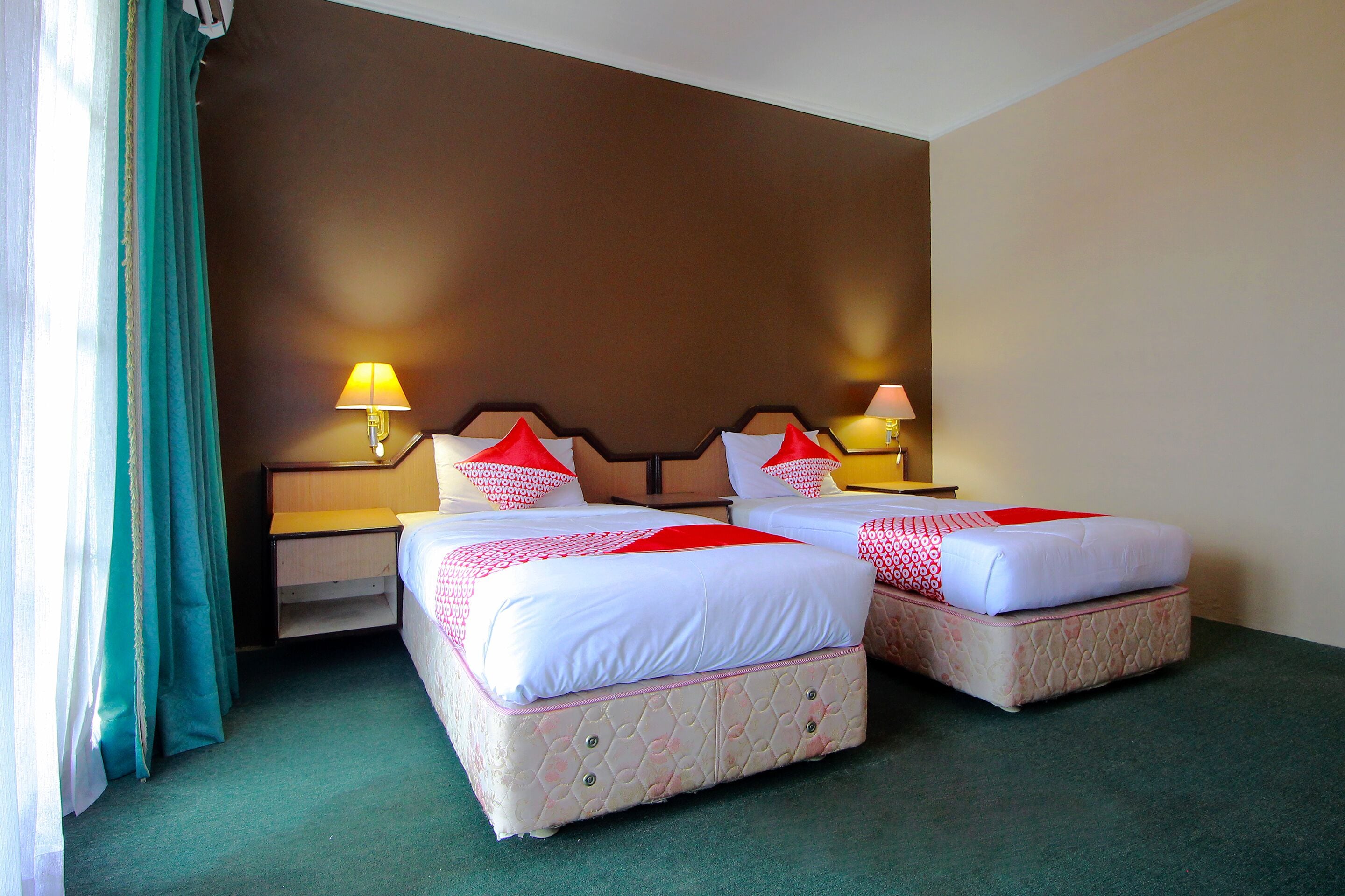A hotel room with two beds, each with a red and white bedspread, two bedside lamps, and a green carpet.