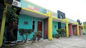 Un bâtiment aux couleurs vives, doté de nombreuses portes et fenêtres, entouré de plantes en pot et d’une cour dallée.