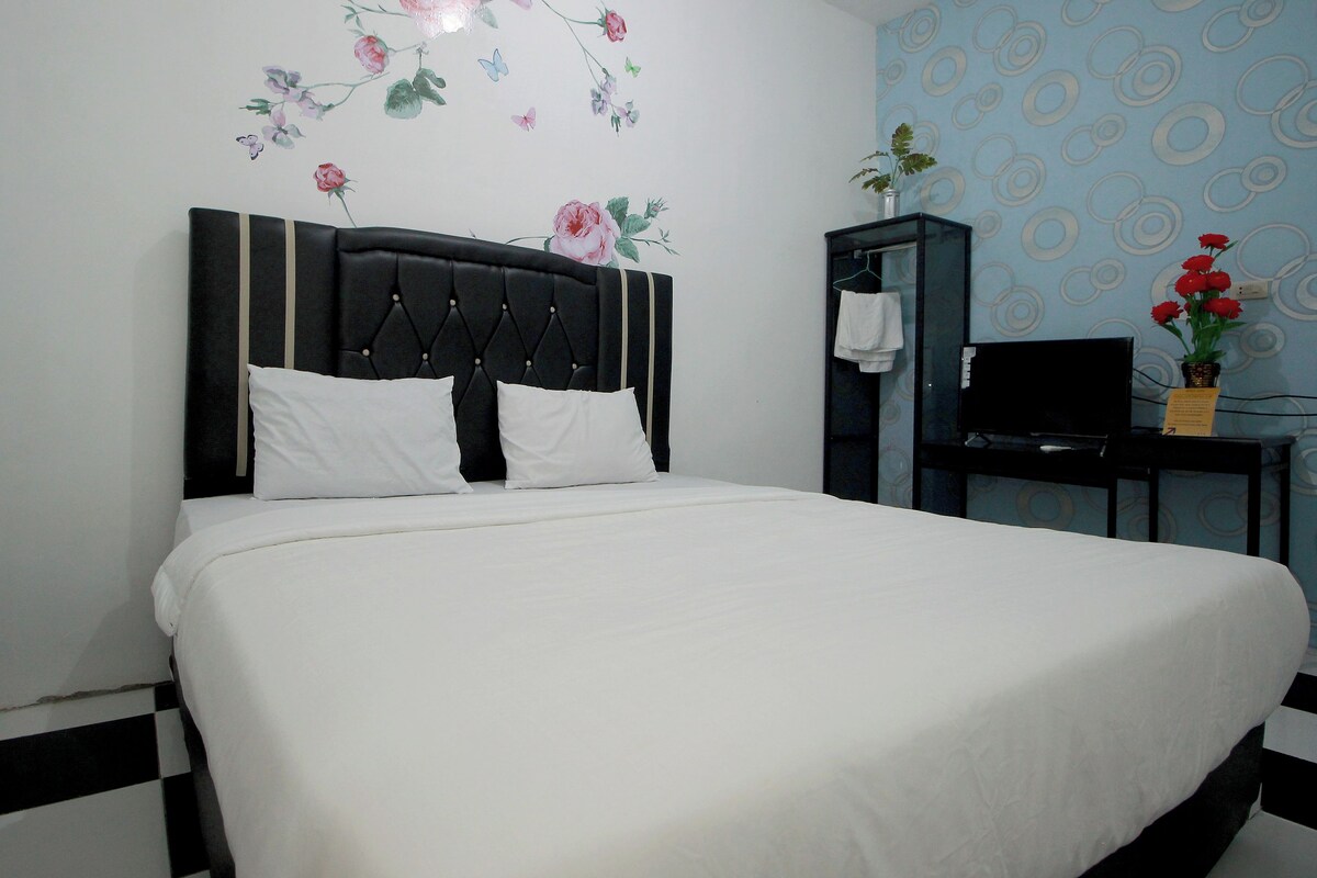 A neatly made bed with a black headboard, a wall with floral decals, a small desk with a TV, and a potted plant with red flowers.