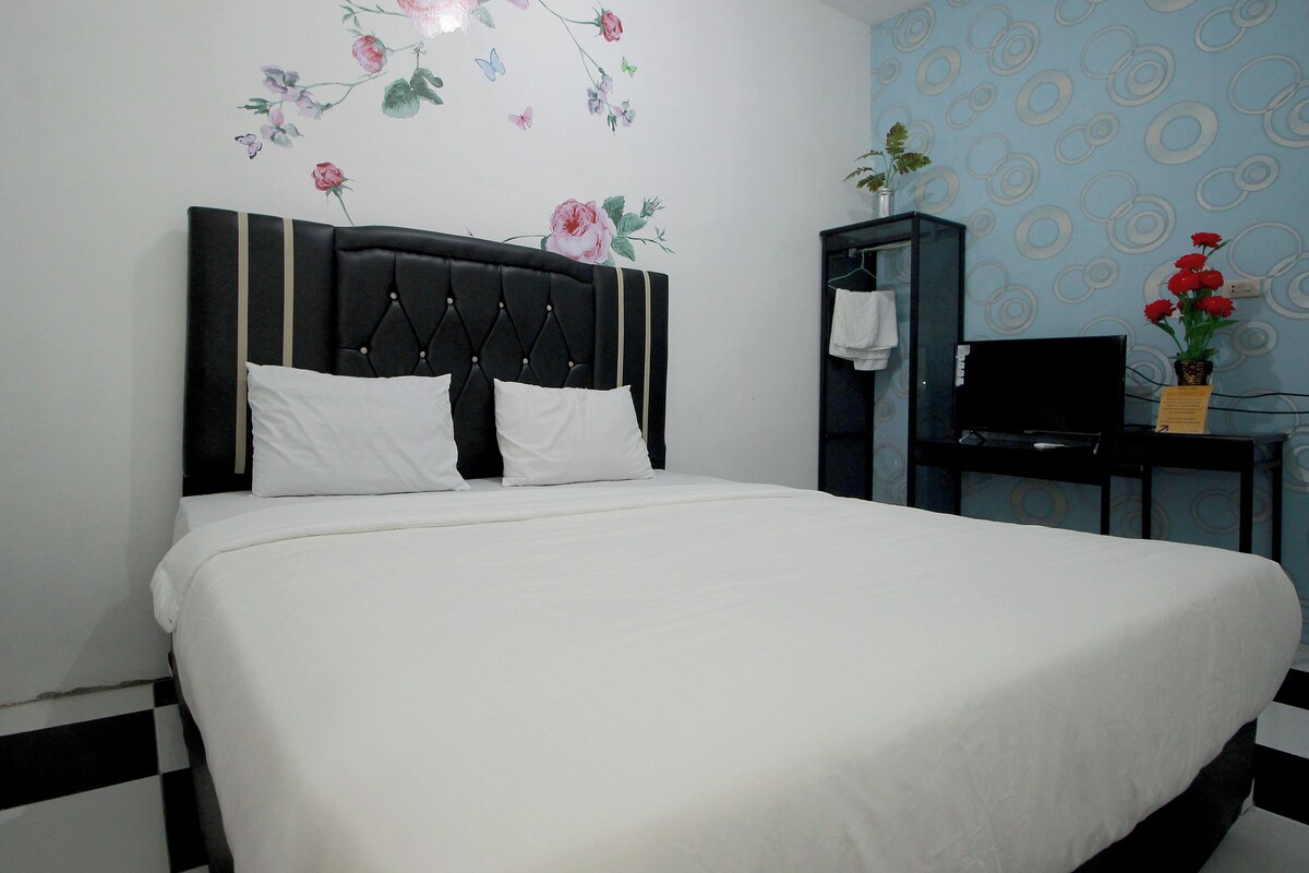 Een netjes opgemaakt bed met een zwart hoofdeinde, een muur met bloemstickers, een klein bureau met een televisie, en een bloempot met rode bloemen.