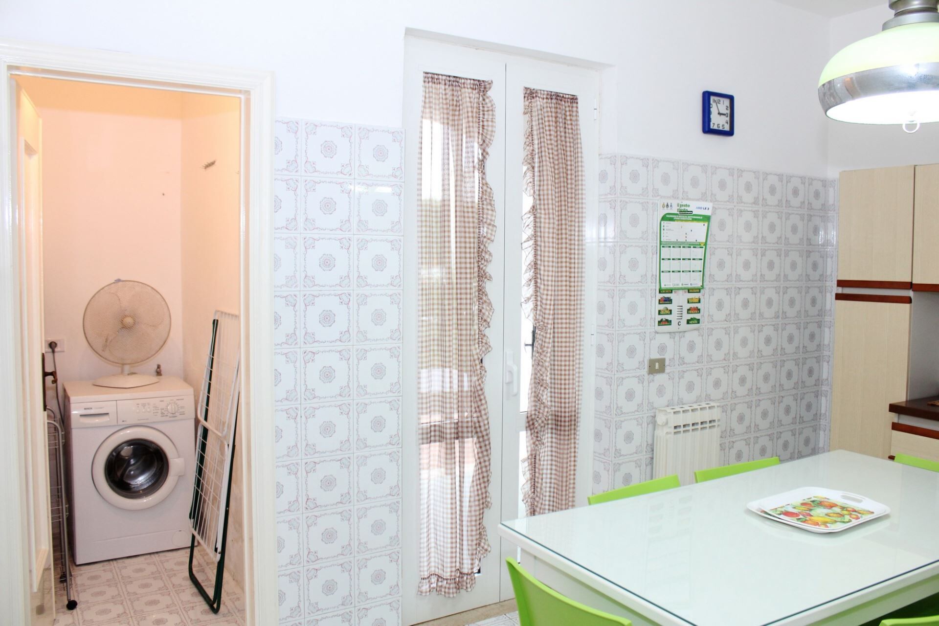 A compact kitchen with a washing machine, a fan, a table with chairs, and a clock on the wall.
