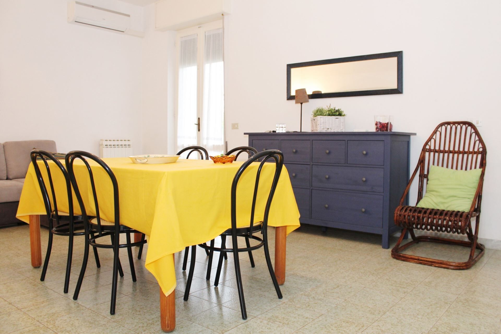 Een eetgedeelte met een gele tafel, vier stoelen, een blauwe kast en een rieten stoel.