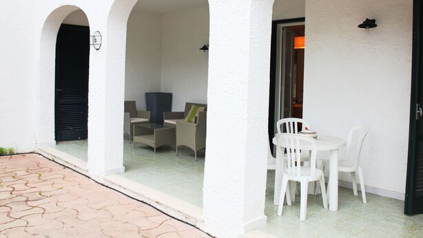 A covered outdoor seating area with white chairs and a table, white walls, and a tiled floor.