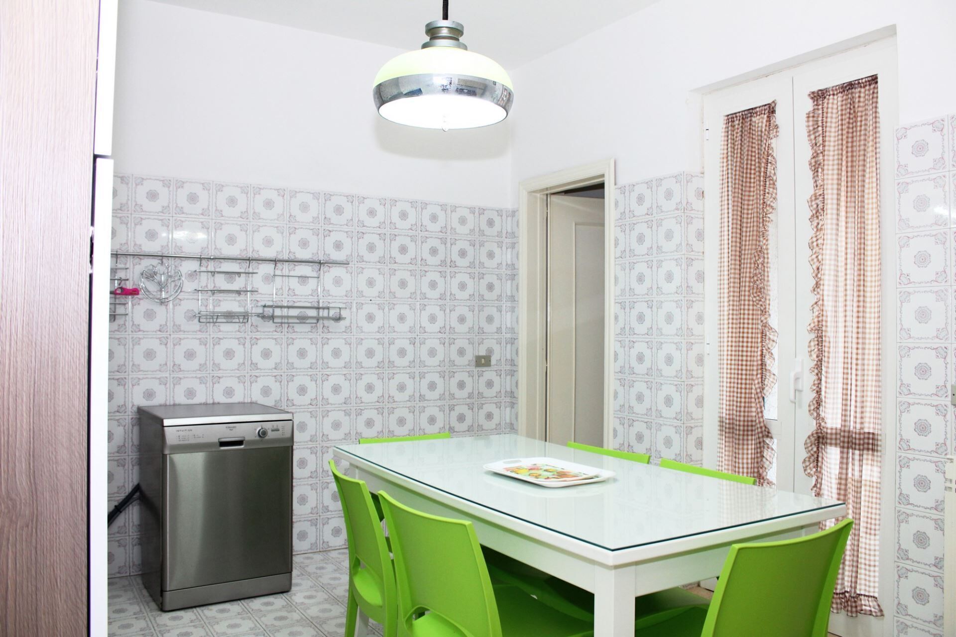 Una cocina compacta con una mesa de vidrio, sillas verdes, lavavajillas de acero inoxidable y papel tapiz estampado.
