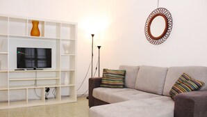 A living room with a sofa, a TV on a stand, a white shelving unit, and a wall-mounted mirror.
