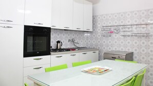 A modern kitchen with white cabinets, a black oven, a gas stove, a kettle, and a small dishwasher. There is a glass table with a green chair and a plate with food.