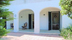 A building with two arched doorways leading to interior spaces, a paved walkway, and outdoor seating with chairs.