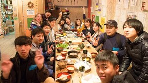 A group of people seated around a table with various dishes, in a room with wooden walls and shelves with books and decorations.