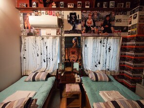 A small, cozy room with two beds, a wooden table, and a wall decorated with various posters and pictures. - Cocoroom Guesthouse Cafe and Garden - Hostel (Osaka)