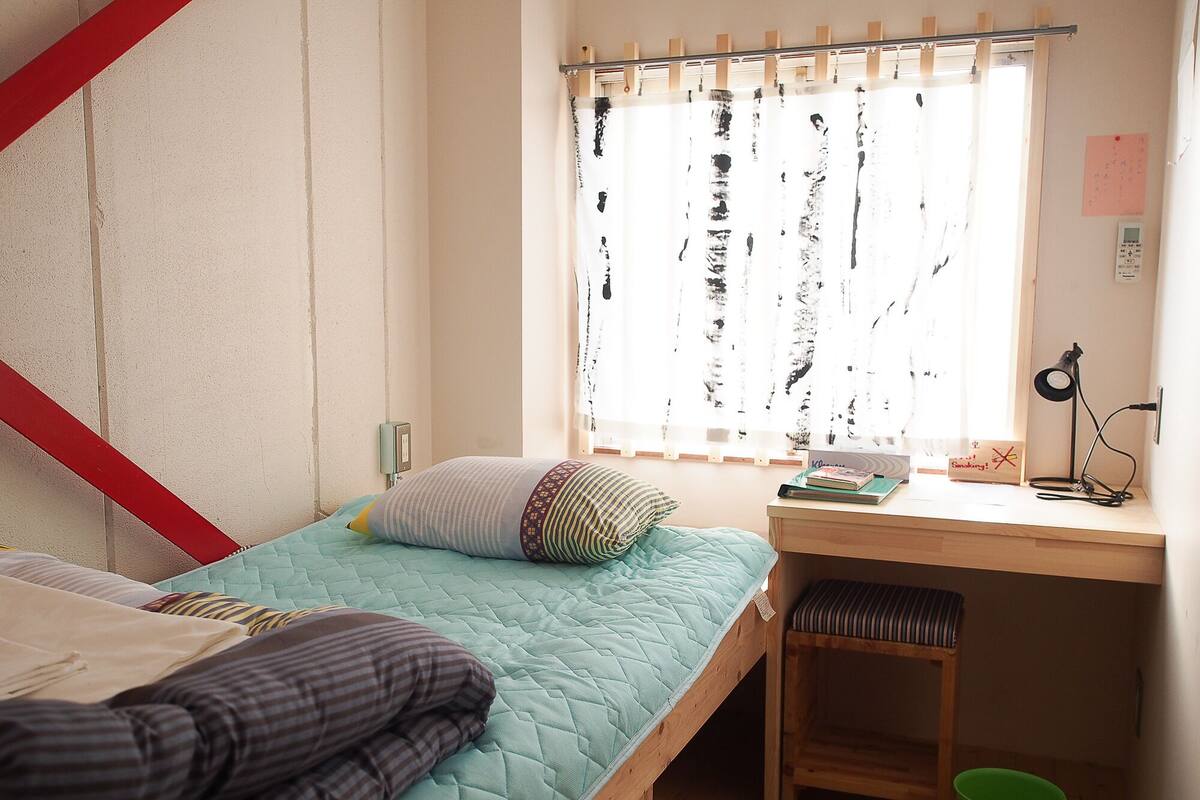 Ein kleines, schlichtes Zimmer mit einem Einzelbett, einem Schreibtisch und einem Fenster mit Vorhang.