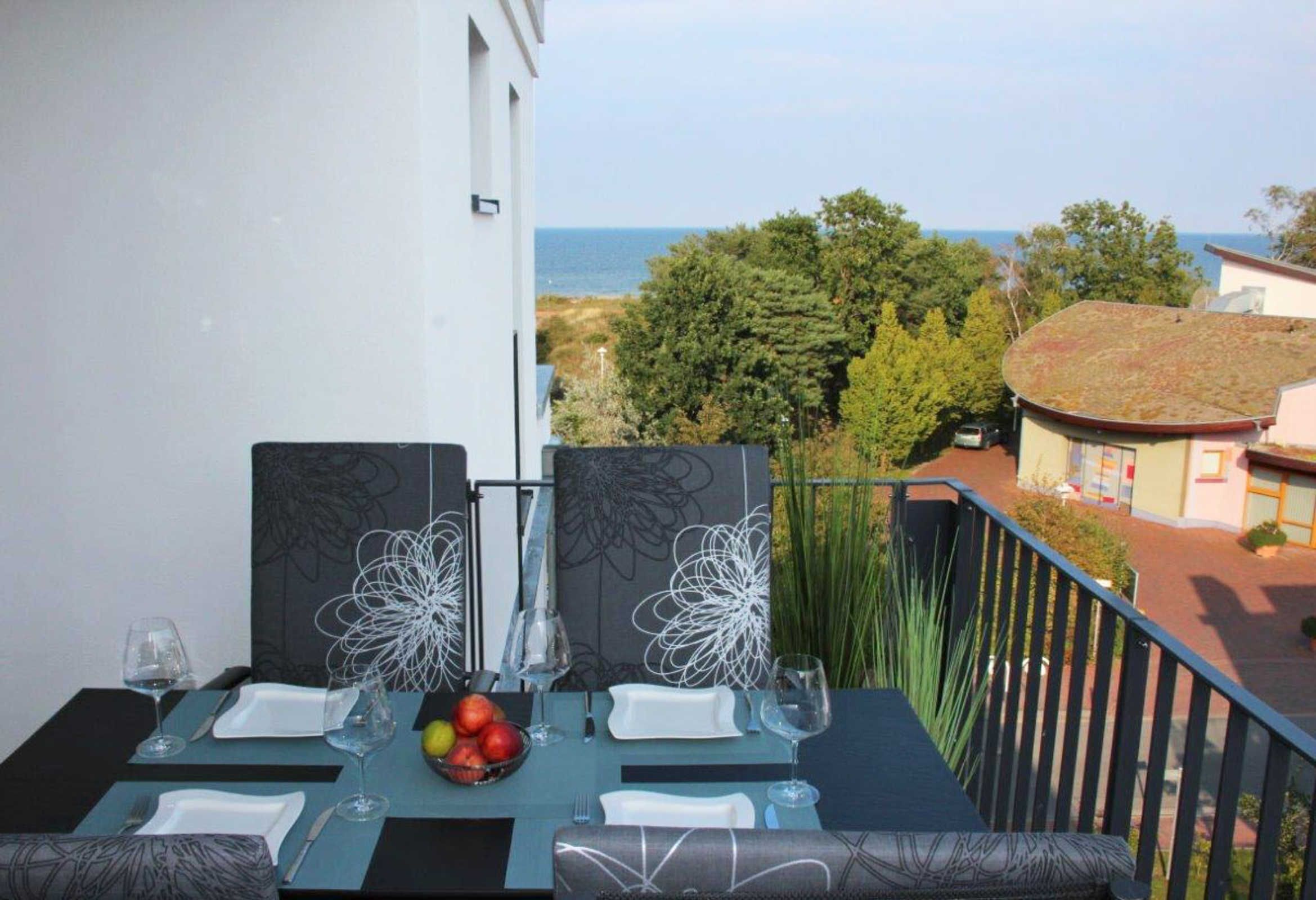 A dining table set with plates, glasses, and a fruit bowl, overlooking a coastal view.