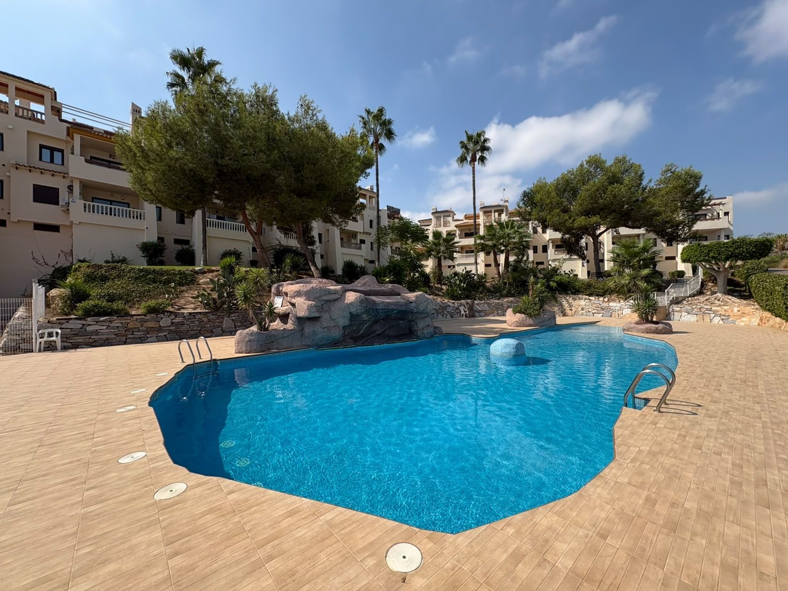 A swimming pool with a clear blue water, surrounded by a tiled deck, with a rocky feature in the middle and trees around.