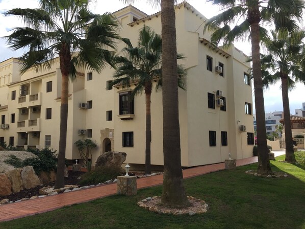 Un edificio de varios pisos con balcones, palmeras y un paseo pavimentado.