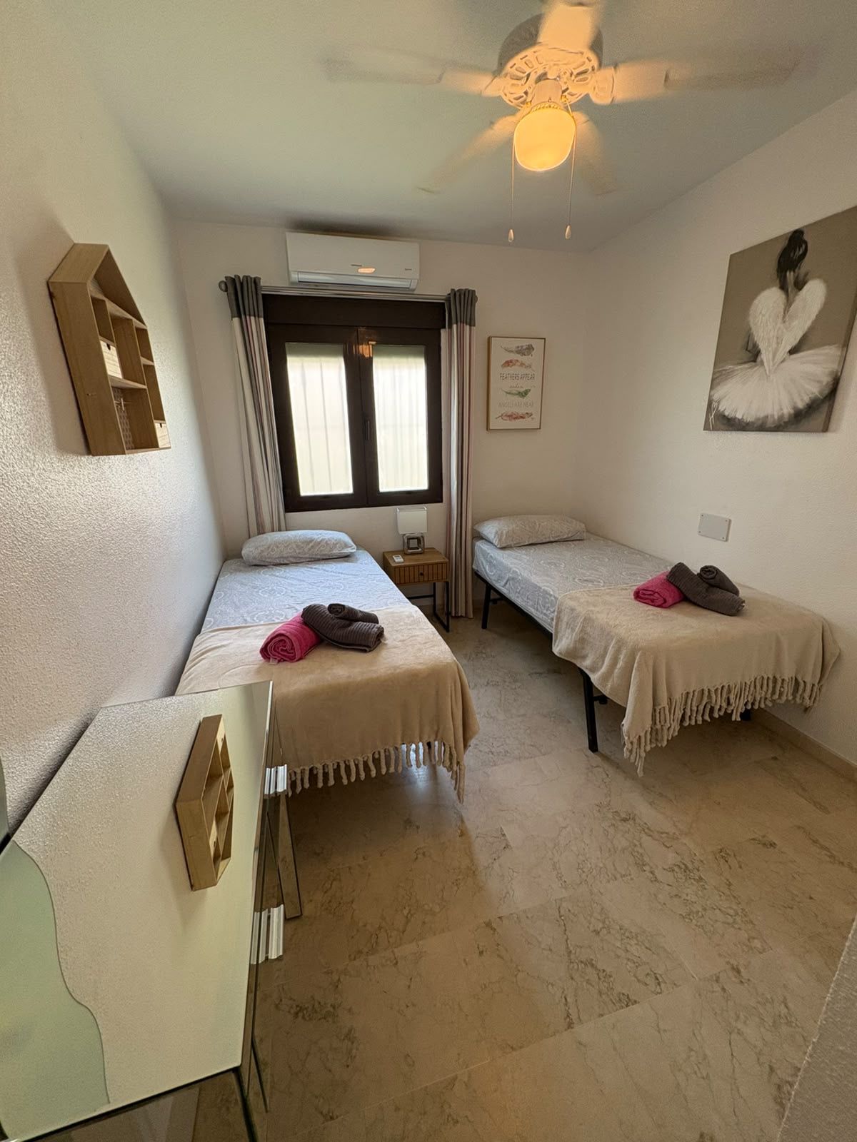 A hotel room with two single beds, a ceiling fan, an air conditioning unit, a small table with a lamp, and a wall-mounted shelf.
