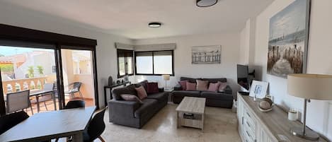 Ein modernes Wohnzimmer mit einer Couch, einem Couchtisch und einem Essbereich. Es gibt Fenster, einen Fernseher und Kunstwerke an den Wänden.
