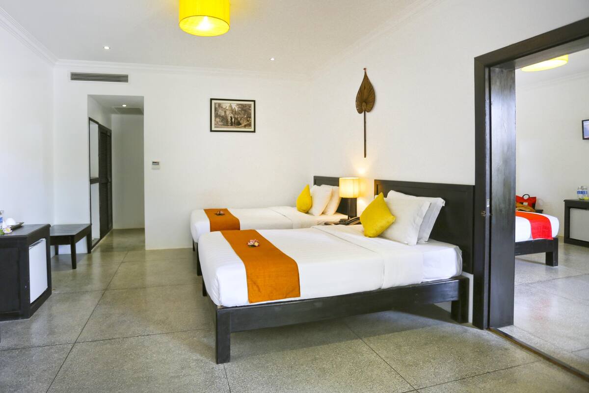 Een hotelkamer met twee bedden, een nachtkastje, een lamp, een foto aan de muur, en een deur naar een andere kamer.