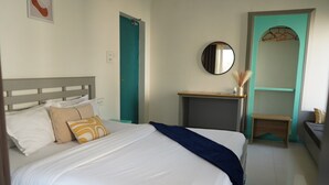 Un lit bien fait, avec du linge de lit blanc et un plaid bleu, une table de chevet avec un miroir, et une porte de couleur turquoise dans une chambre d’hôtel.