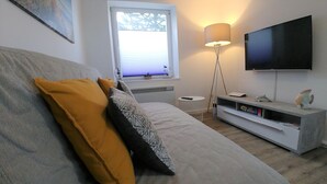 A modern living room with a large sofa, a flat-screen TV mounted on the wall, a floor lamp, and a small side table with a remote control. - Apartment Möwennest Oberhof Boltenhagen (Oberhof)