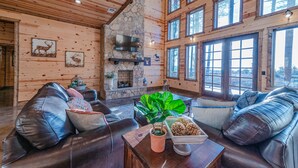 A spacious living room with leather sofas, a stone fireplace, large windows, and wooden walls.