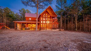 A large wooden cabin with a prominent gable roof, multiple windows, and a gravel driveway.
