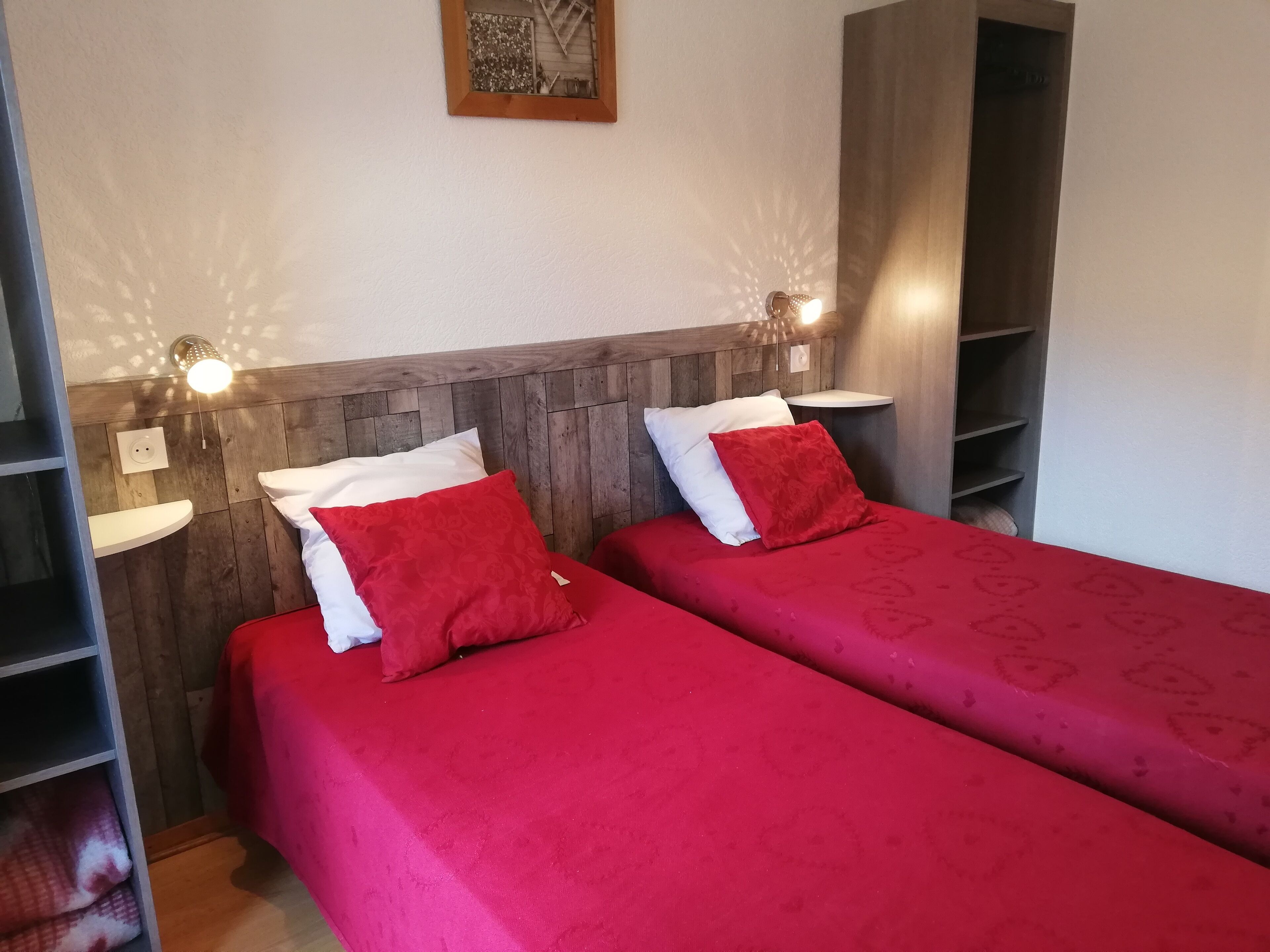 Een kamer met twee bedden, elk met rode beddengoed en witte kussens. Aan weerszijden van de bedden zijn wandlampen bevestigd, en er is een houten hoofdbord.