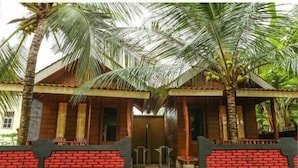 A wooden house with a brick facade, surrounded by palm trees and greenery. - Watukarung Prapto Homestay (Sendang)