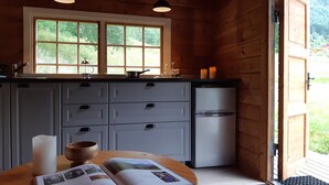 Cocina de cabaña de madera con un ventanal grande, un refrigerador y una mesa con un libro y una vela.